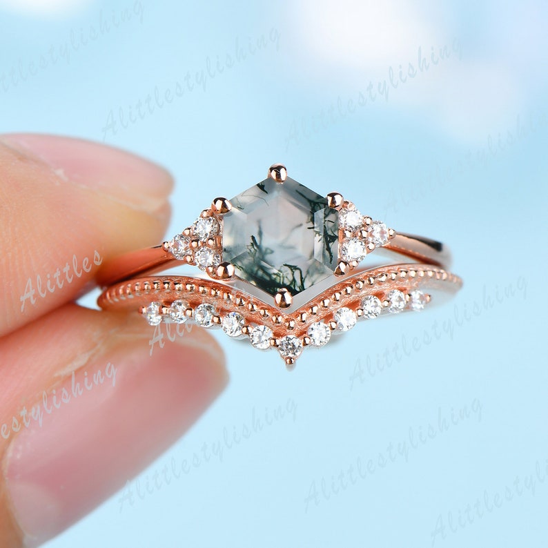 May include: Rose gold engagement ring set with a hexagonal moss agate center stone and a delicate pave band. The ring is accented with small clear cubic zirconia stones.