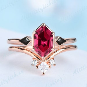 May include: A rose gold engagement ring set with a large octagon-shaped ruby gemstone, two smaller black gemstones, and smaller white gemstones. The ring is designed with a V-shaped band.