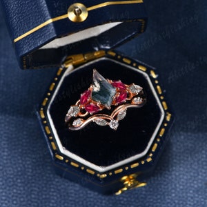 May include: A rose gold ring set with a green and black gemstone surrounded by red ruby stones. The ring is in a blue velvet ring box.