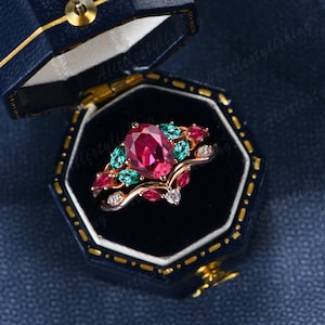 May include: A rose gold ring set with a large oval ruby surrounded by smaller emerald and ruby stones. The ring is in a blue velvet ring box.