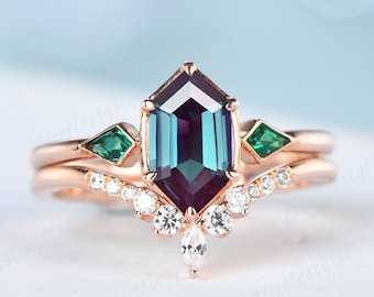 May include: A rose gold engagement ring set with a large octagon-shaped alexandrite gemstone, surrounded by smaller diamonds. The ring also features two emerald green accent stones.