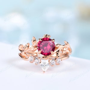 以下が含まれることがあります： 大きな丸い赤い宝石をあしらった、ローズゴールドの婚約指輪。葉のデザインが特徴で、小さな白い宝石が付いたお揃いのバンドが付いています。指輪は柔らかくぼやけた背景にセットされています。