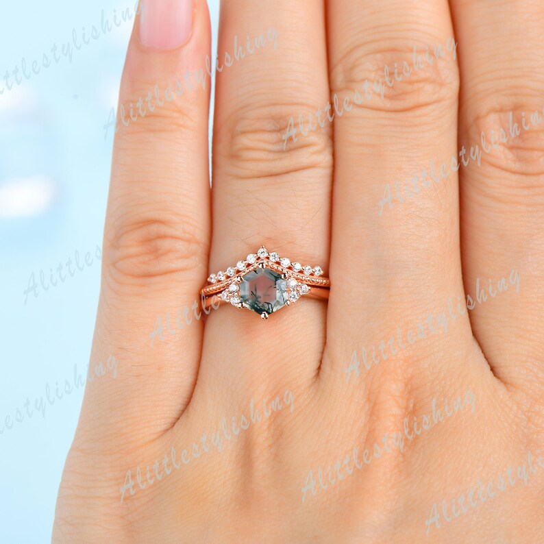 May include: A rose gold engagement ring set with a hexagonal green gemstone surrounded by smaller clear gemstones. The ring is on a finger.