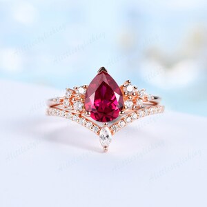 May include: A pear-shaped ruby engagement ring set in rose gold with a matching band featuring a chevron design and smaller diamonds.