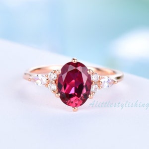 May include: A rose gold ring featuring a large oval ruby gemstone surrounded by smaller white gemstones. The ring has a delicate design with two pear-shaped white gemstones on either side of the ruby.
