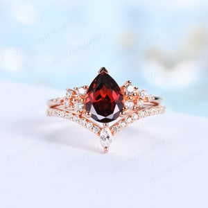 May include: A pear-shaped garnet engagement ring set in rose gold with a delicate diamond halo and a matching diamond band. The ring features a v-shaped band with pave diamonds.