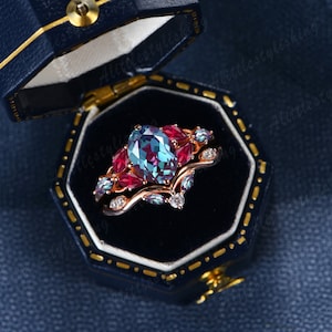May include: A rose gold ring set with a large oval alexandrite gemstone surrounded by smaller red and blue gemstones. The ring is in a blue velvet ring box.