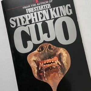 May include: A paperback book titled "CUJO" by Stephen King. The cover features a menacing dog's face with exposed teeth against a black background. The book is from the author of "Firestarter".