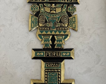 May include: A gold-colored metal wall hanging featuring an Incan figure with intricate details. The figure has a headdress and is adorned with turquoise accents. The word "PERU" is displayed, along with "SIPAN".