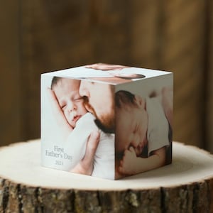 Op de afbeelding: Een kubusvormig fotoblok met afbeeldingen van een baby en een persoon. Het blok heeft de tekst "First Father's Day 2024" erop. Het blok wordt gepresenteerd op een houten oppervlak.