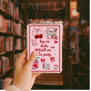 Peut inclure: Une coque de téléphone transparente avec un motif vichy rose et divers autocollants. Les autocollants incluent des cerises, un cocktail, un livre et le texte "buy me books & tell me I'm pretty". La coque est tenue dans une main.