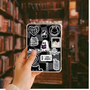 May include: A clear phone case with a black and white checkered design. The case is decorated with various stickers, including a ghost reading a book, cherries, and the words "Bestie Vibes Only" and "Dark Romance".