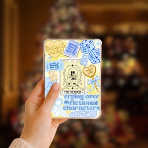 May include: A clear phone case with a yellow background and various stickers. The stickers include text that says "Rather be reading", "The Reader", and "crying over fictional characters". Other stickers include a ghost, a teddy bear, and a heart-shaped lollipop.