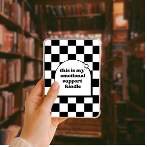 Könnte beinhalten: Kindle-Hülle mit einem schwarz-weißen Schachbrettmuster. Die Hülle hat eine weiße Sprechblase mit dem Text "this is my emotional support kindle" und einen kleinen Stern. Der Hintergrund ist ein verschwommenes Bild eines Bücherregals.