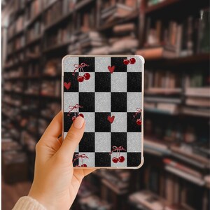 May include: A clear phone case with a black and silver checkered pattern. The case is decorated with red cherries and hearts. The phone case is held in a person's hand.