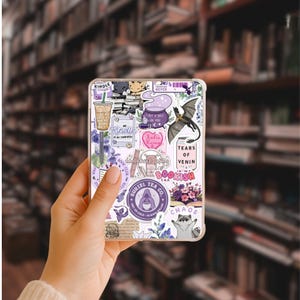 May include: A clear Kindle case decorated with stickers. The stickers include a purple coffee cup, a dragon, and the words "Kindle," "Bookish," and "Chaos." The case is held in a person's hand against a blurred bookshelf background.
