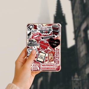 May include: Clear phone case with a collage of stickers. Stickers include text like "IS THAT SMUT?" and "MAFIA ROMANCE", along with graphic elements such as a heart, lips, and a chili pepper. The case is held in a hand.