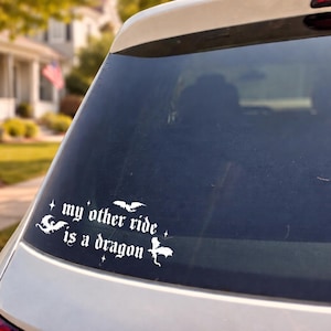 May include: White vinyl decal applied to a car window. The decal features the text "my other ride is a dragon" in a gothic font, along with illustrations of a dragon, bats, and stars. The car is a light color, and the background is blurred.