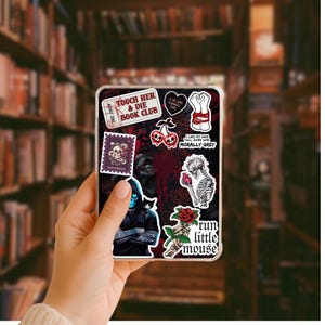 May include: A clear phone case with a dark, gothic design. The case features various stickers with text like "Touch Her & Die Book Club" and illustrations of skulls, roses, and a skeleton. The background is a blurred image of a bookshelf.