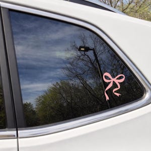 May include: A pink bow decal is affixed to the window of a white vehicle. The bow is a simple design with two loops and two flowing tails. The background shows a reflection of trees and a cloudy sky.