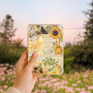 May include: A clear phone case featuring a collage of vintage botanical illustrations. The design includes sunflowers, butterflies, and a yellow bow on a cream-colored background. The case is held in a hand against a blurred outdoor backdrop.