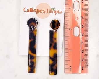 May include: A pair of tortoiseshell earrings with a round top and a long rectangular bottom. The earrings are on a white card with the text "Calliope's Utopia".