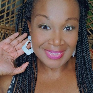 May include: A woman with black braided hair wearing large white geometric earrings. She is smiling and looking at the camera.