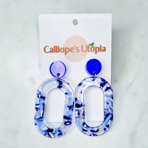 Könnte beinhalten: Ein Paar blaue und weiße ovale Ohrringe mit einem blauen Kreis an der Oberseite jedes Ohrrings. Die Ohrringe befinden sich auf einer weißen Karte mit dem Text "Calliope's Utopia" in Orange.