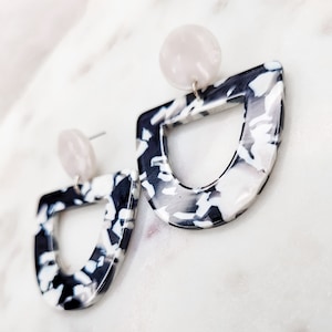 Puede incluir: Un par de pendientes con estampado en blanco y negro con un acento redondo blanco en la parte superior. Los pendientes tienen forma de semicírculo con un recorte en el medio.