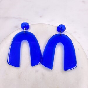 Peut inclure: Une paire de boucles d'oreilles en acrylique bleu en forme d'arche. Les boucles d'oreilles sont une pièce audacieuse et moderne.