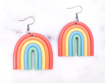 Könnte beinhalten: Ein Paar regenbogenfarbene Ohrringe in Form eines Regenbogenbogens. Die Ohrringe sind aus Polymer Clay gefertigt und haben einen silbernen Haken.
