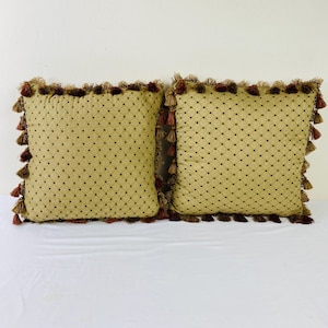 May include: Two square throw pillows in a muted gold fabric featuring a dark brown diamond grid pattern. Each pillow is finished with a decorative fringe border composed of small, hanging tassels in alternating shades of brown and gold.