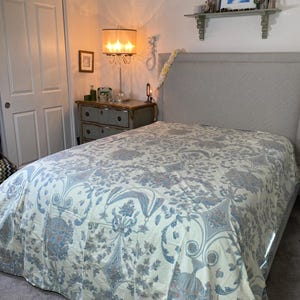 May include: A bed with a light gray headboard and a floral patterned bedspread. The bedspread features a cream background with blue and gray floral designs. A bedside table with a lamp and a framed picture are visible.