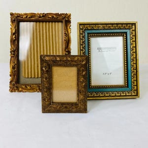 May include: Three rectangular picture frames. One large frame has an ornate gold finish and a corrugated cardboard insert. A smaller, dark bronze-toned frame sits in front. A third frame features a gold outer border, a teal inner border, and a 3.5" x 5" label.
