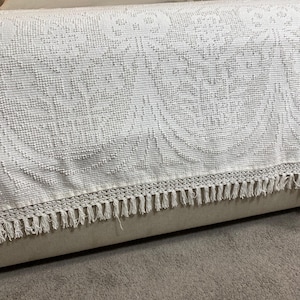 May include: A white bedspread featuring a raised, textured floral pattern across the fabric. The bottom edge is finished with a decorative crocheted border and a row of long, thin, hanging fringe tassels that drape over the side of the bed.
