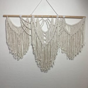 May include: A cream-colored macrame wall hanging. The piece is suspended from a natural wood dowel and features intricate knotting patterns. The design includes three distinct sections with cascading fringe, creating a textured, bohemian aesthetic.