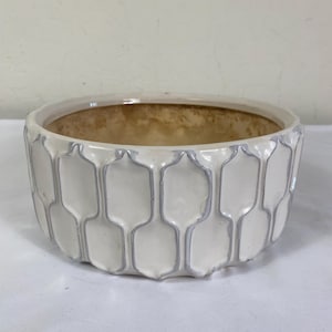 May include: A white ceramic planter with a geometric pattern and a light brown interior. The planter has a wide, shallow bowl shape and is suitable for indoor or outdoor use. The planter's dimensions are approximately 10 inches in diameter and 5 inches tall.