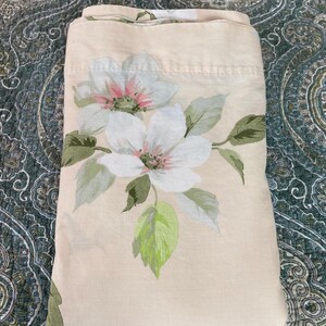 May include: A folded cream-colored bed sheet featuring a floral pattern. The design includes large white flowers with pink centers and green leaves. The sheet is made of a soft fabric and is folded neatly.