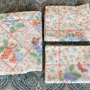 May include: A set of three white fabric pieces featuring a repeating pattern of orange and blue flowers, green leaves, and butterflies. The design is divided by thin red grid lines, creating a soft floral aesthetic across the textile surfaces.