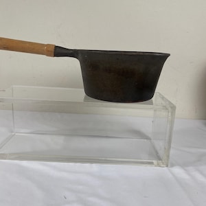 May include: A small, dark-colored cast iron saucepan with a wooden handle. The pan has a rounded shape and a single, long handle extending from the side. The handle is made of light-colored wood. The pan is sitting on a clear acrylic surface.