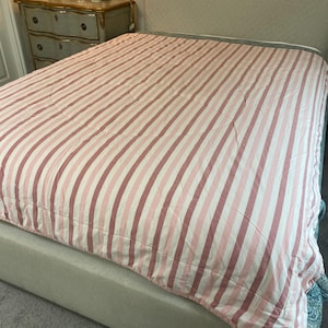 May include: A bed covered with a pink and white striped comforter. The comforter has a circular quilted pattern in the center. The bed frame is a light beige color. A dresser and lamp are visible in the background.