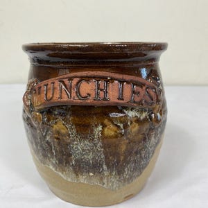 May include: A handcrafted ceramic jar with a brown glaze and the word "MUNCHIES" in raised letters. The jar has a textured surface and a light tan base. The jar is suitable for kitchen storage.