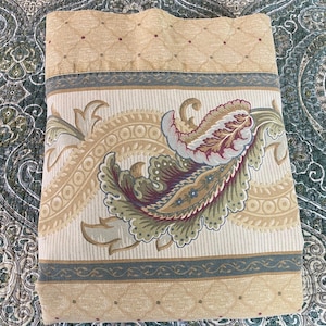 May include: A folded beige bed sheet with a detailed paisley design. The sheet features a border with a repeating pattern of small diamonds and a central band with a large paisley motif in shades of green, burgundy, and beige.