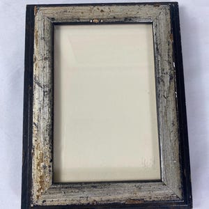 May include: Rectangular picture frame with a distressed silver and black finish. The frame has a rectangular opening with a cream-colored backing. The frame's edges show signs of wear, adding to its vintage aesthetic.