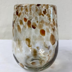 May include: A clear, stemless glass with a speckled pattern of brown and tan spots. The glass has a rounded shape and a thick base. The speckled design covers the majority of the glass, creating a unique visual texture.