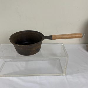 May include: A small, dark brown cast iron saucepan with a long, light-colored wooden handle. The pan has a rounded shape and a single handle. The handle is attached with a metal ring. The pan is sitting on a clear acrylic surface.