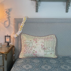 May include: A floral patchwork pillow with a pink border rests on a bed with a patterned blue comforter. A white flower garland hangs from the headboard. A small lamp and a decorative shelf are visible in the background.