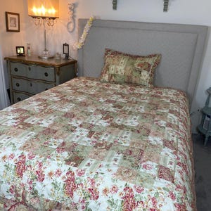 May include: A patchwork quilt and matching pillow sham featuring a floral pattern in shades of sage green, cream, and deep red. The bedding displays a repeating square block design with intricate botanical prints and a ruffled edge along the bottom.