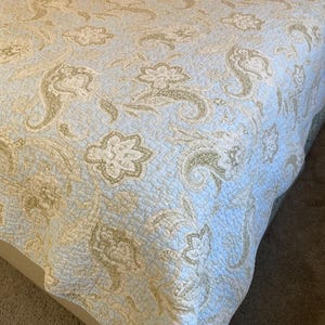 May include: A light blue quilted bedspread with a floral and paisley pattern in shades of beige and cream. The bedding has a stitched, textured surface and is draped over a bed.
