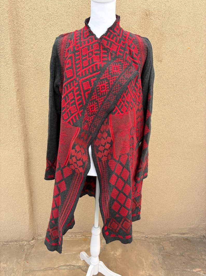 Puede incluir: Un c&aacute;rdigan largo y abierto en gris y rojo, exhibido en un maniqu&iacute; blanco. El c&aacute;rdigan presenta un patr&oacute;n geom&eacute;trico en rojo sobre un fondo gris. Las mangas son grises lisas.